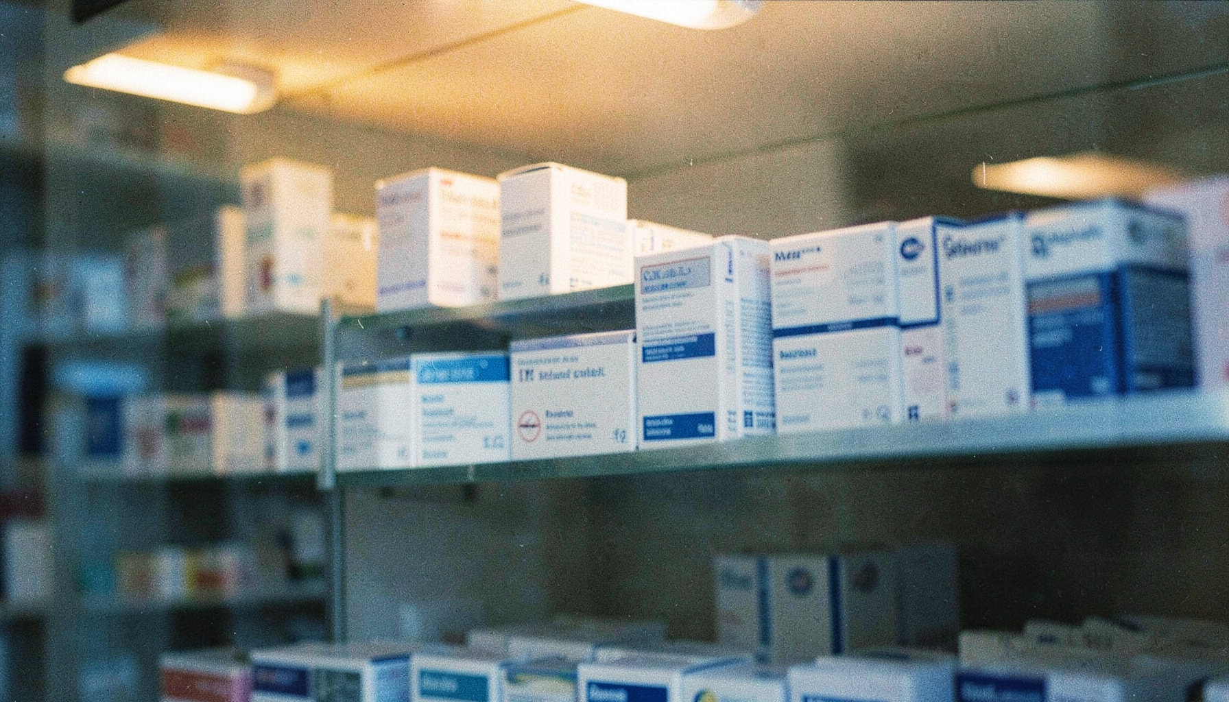 A softly lit pharmacy shelf with neatly arranged white and blue medicine boxes under warm amber overhead lighting